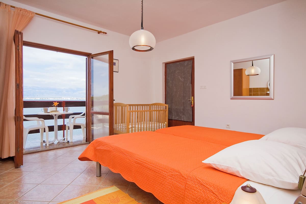 A bedroom with a large bed, a crib, a balcony with a table and chairs, and a view of the sea.