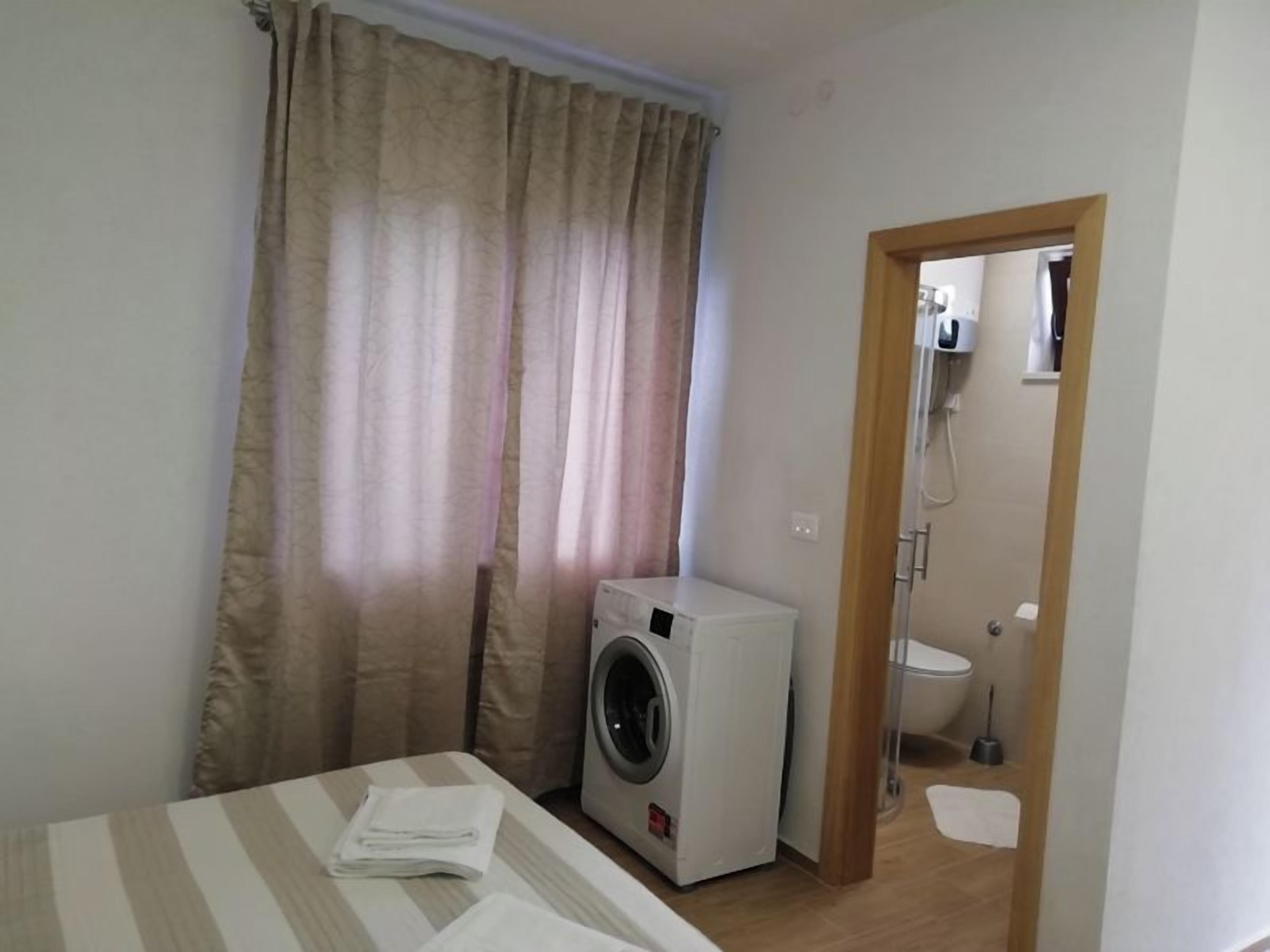 A small room with a washing machine, a bed with a striped bedspread, and a door leading to a bathroom.
