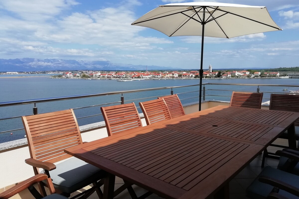 A wooden table and chairs with a white umbrella on a balcony overlooking a body of water and a town.