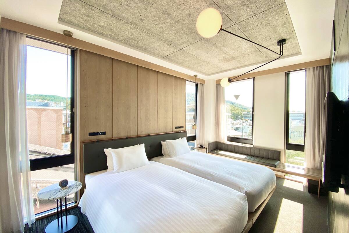 Habitación de hotel con dos camas, una mesita pequeña, un sofá y ventanas amplias con vistas a la ciudad.