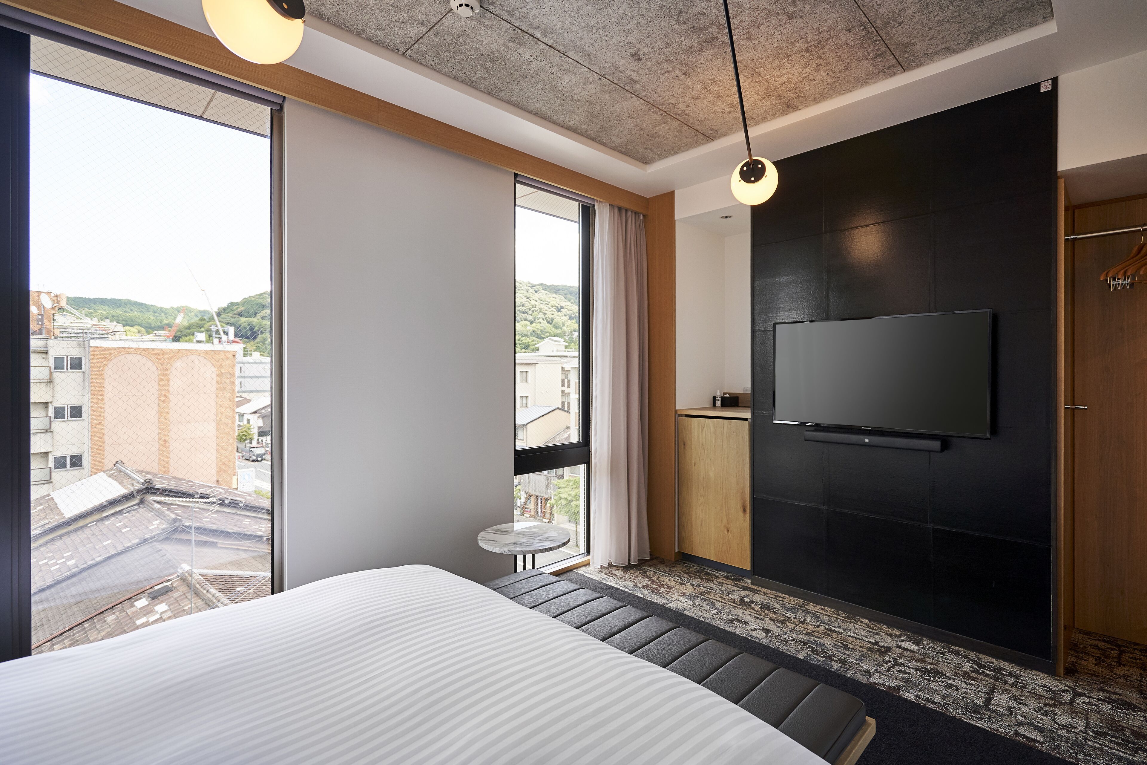 Une chambre d’hôtel moderne dotée d’un grand lit, d’un téléviseur fixé au mur, d’une petite table et offrant une vue sur la ville grâce à de grandes fenêtres.