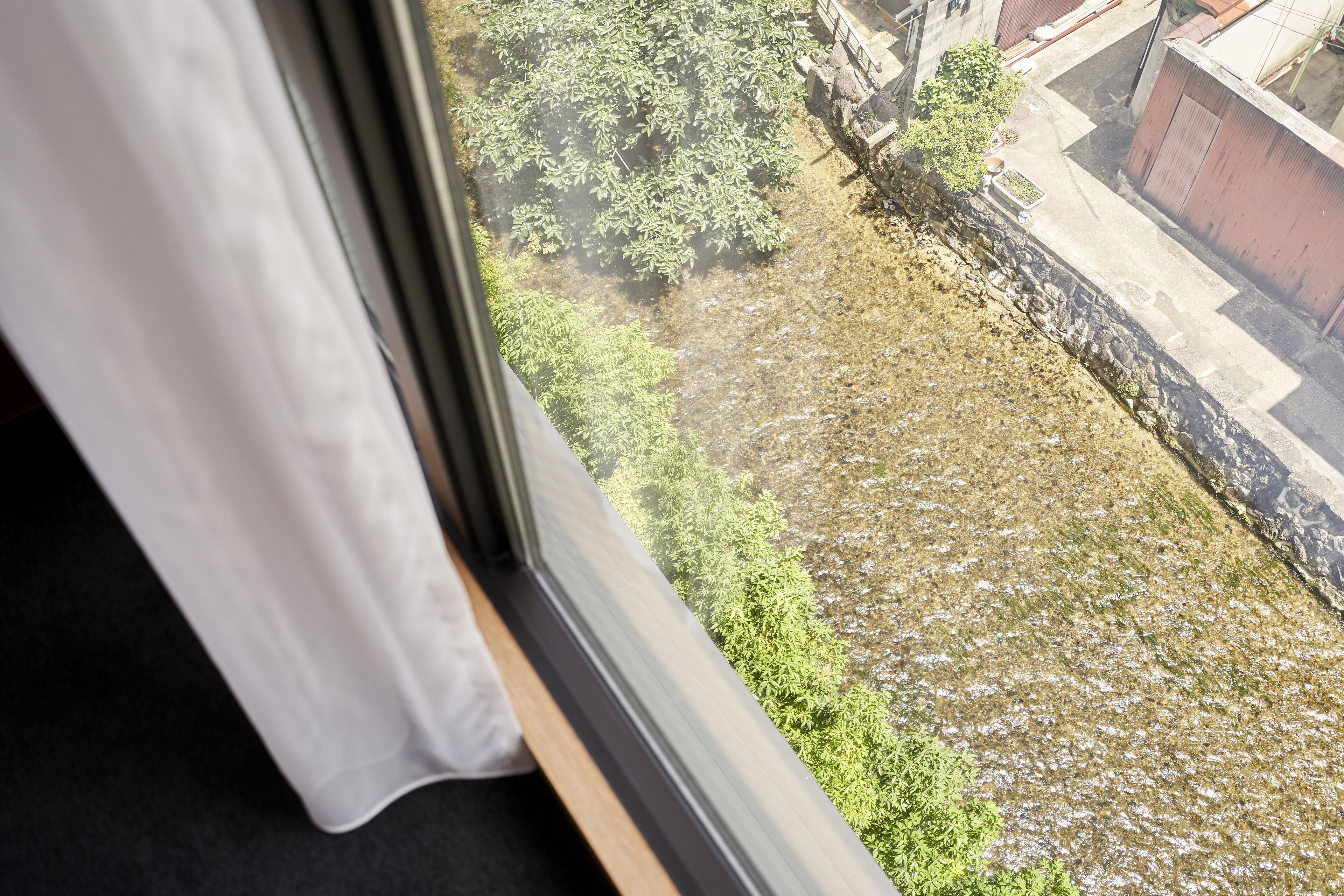 Näköala ikkunasta, jonka edessä on verho. Näkymä avautuu sisäpihalle, jossa on vihreää kasvillisuutta ja kivimuuri.