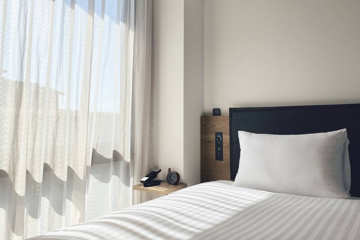 Una habitación de hotel con una cama, una mesita de noche con un reloj, un enchufe y cortinas translúcidas.