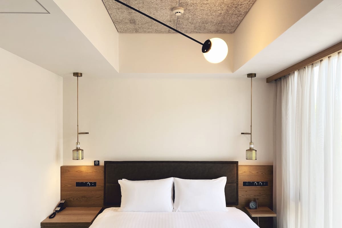 Una habitación de hotel moderna con una cama grande, cabecera de madera, dos mesitas de noche con lámparas y un techo con textura.