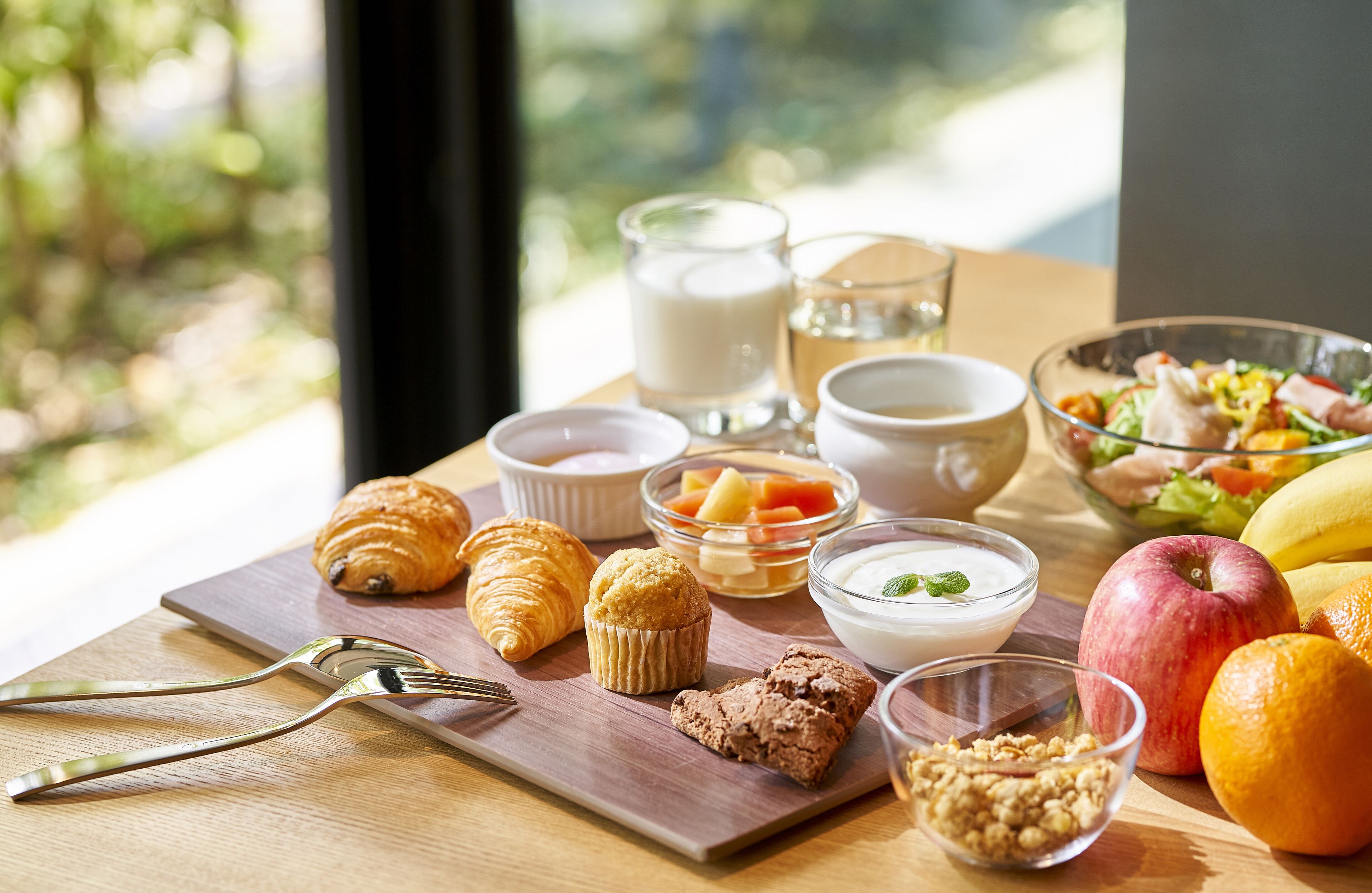 A breakfast table with croissants, muffins, yogurt, fruit, and a bowl of salad.
