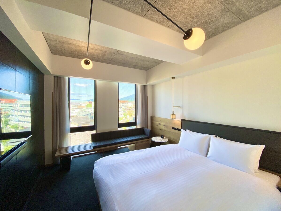 A hotel room with a large bed, a bench by the window, and a view of the city.