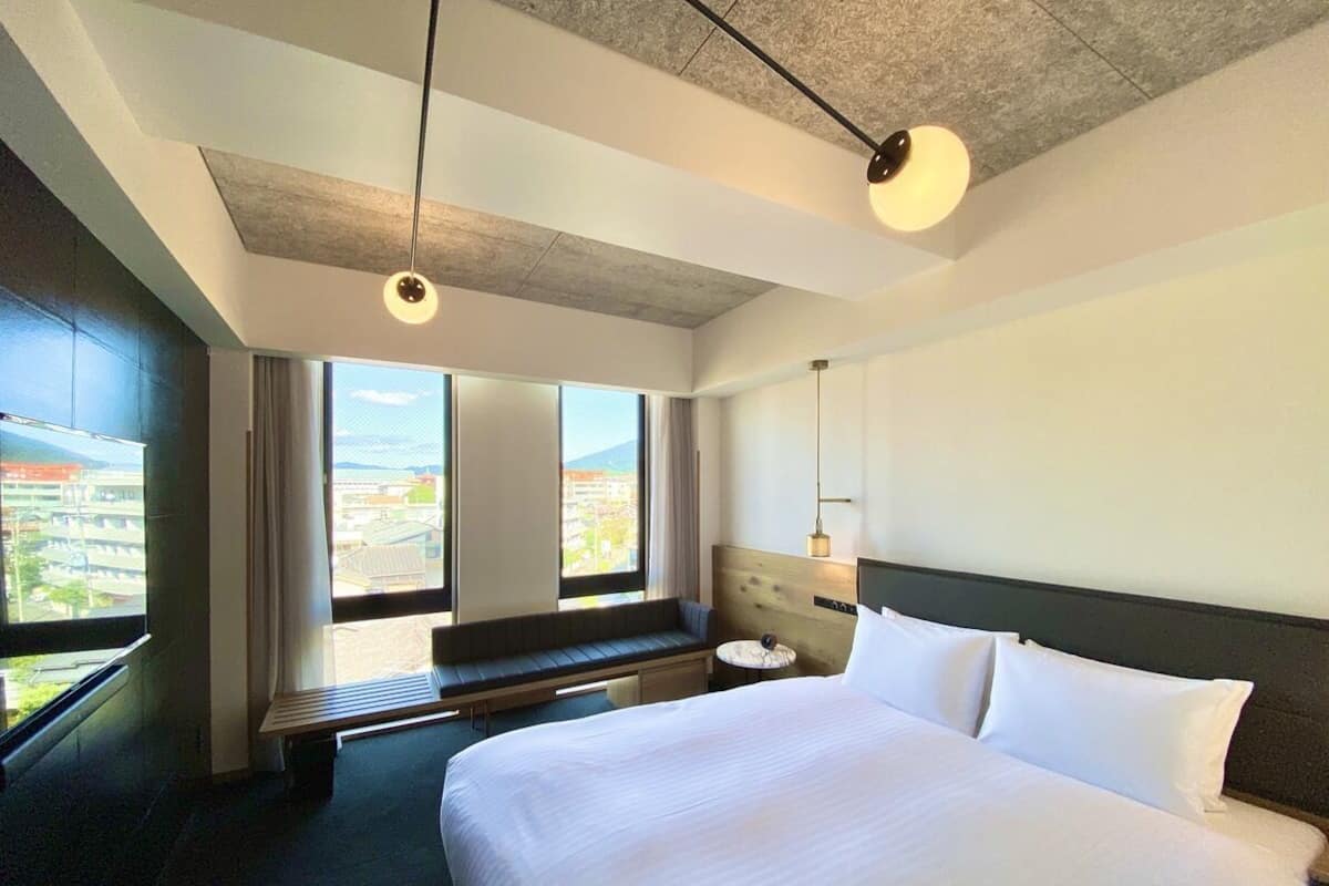 Habitación de hotel con una cama grande, un banco junto a la ventana y vistas a la ciudad.
