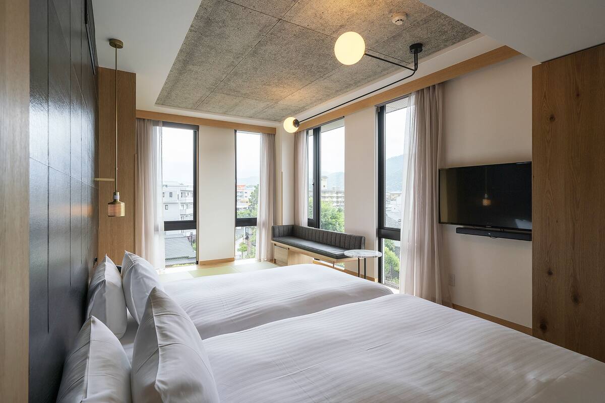 Una habitación de hotel moderna con una cama grande, un televisor montado en la pared y vistas a la ciudad a través de amplias ventanas.