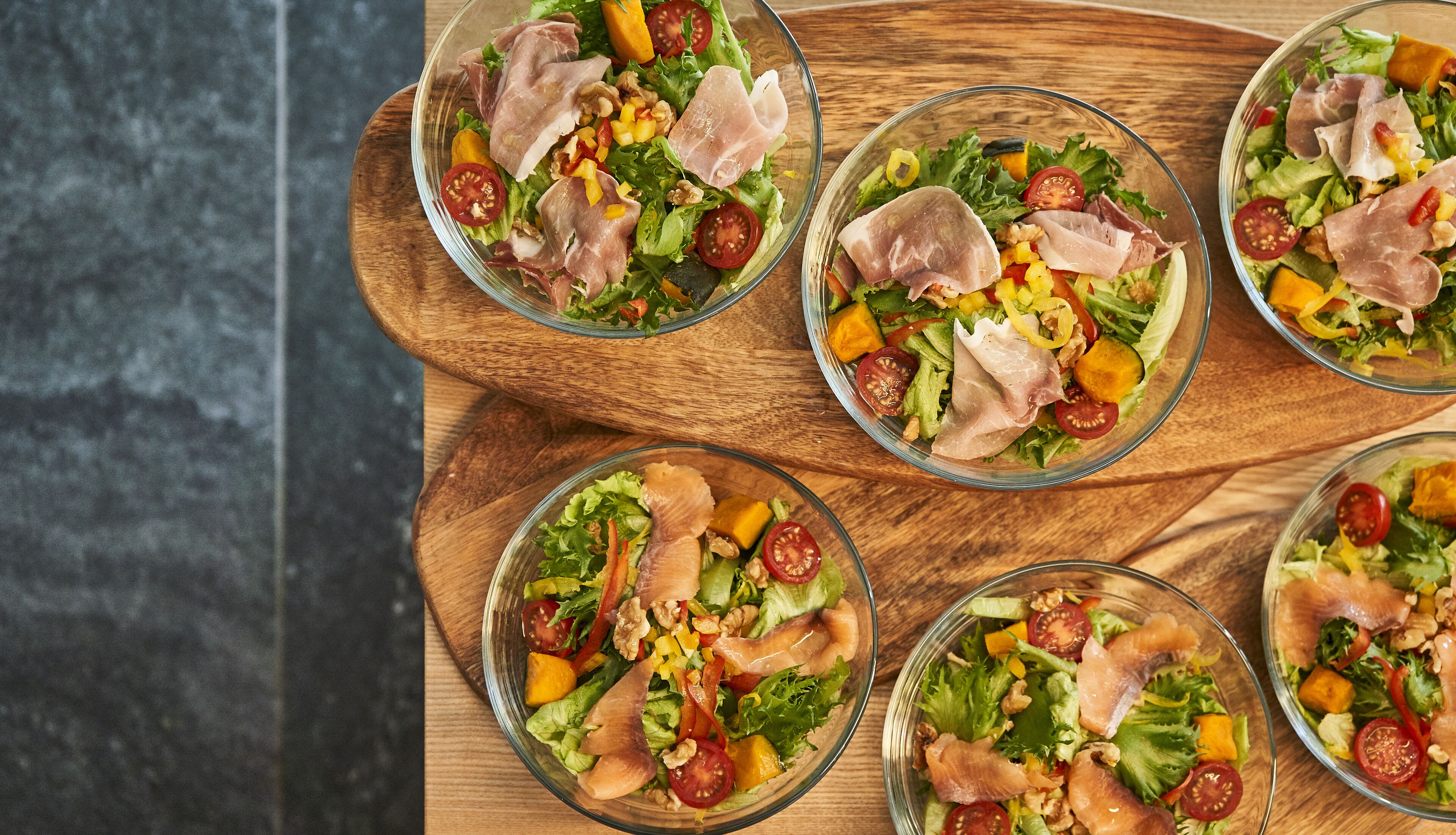 A wooden table with five glass bowls of salad containing lettuce, tomatoes, corn, and slices of meat.