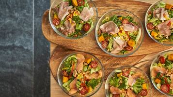 A wooden table with five glass bowls of salad containing lettuce, tomatoes, corn, and slices of meat.