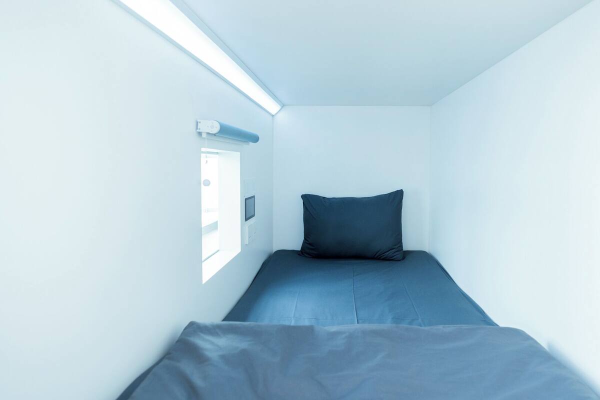 A small, narrow room with a single bed, a window, and a wall-mounted control panel.