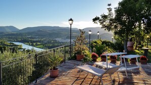 Una terraza con vistas a un río y montañas, amueblada con sillones blancos y plantas en macetas.