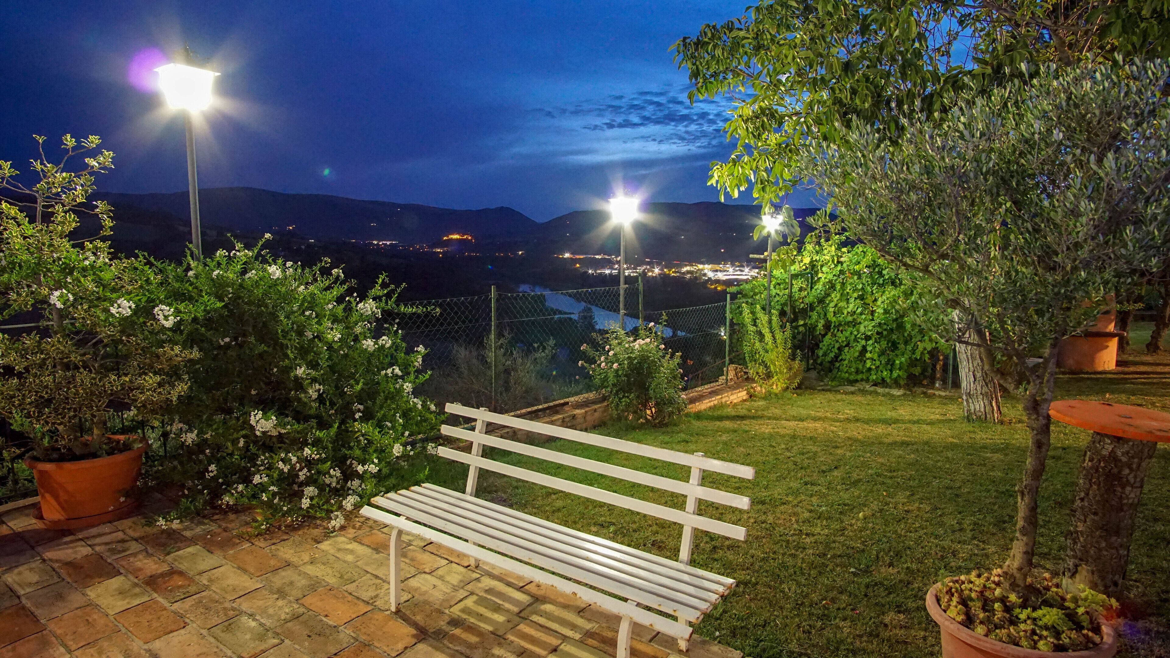 An outdoor patio with a white bench, potted plants, and a view of a cityscape at night.