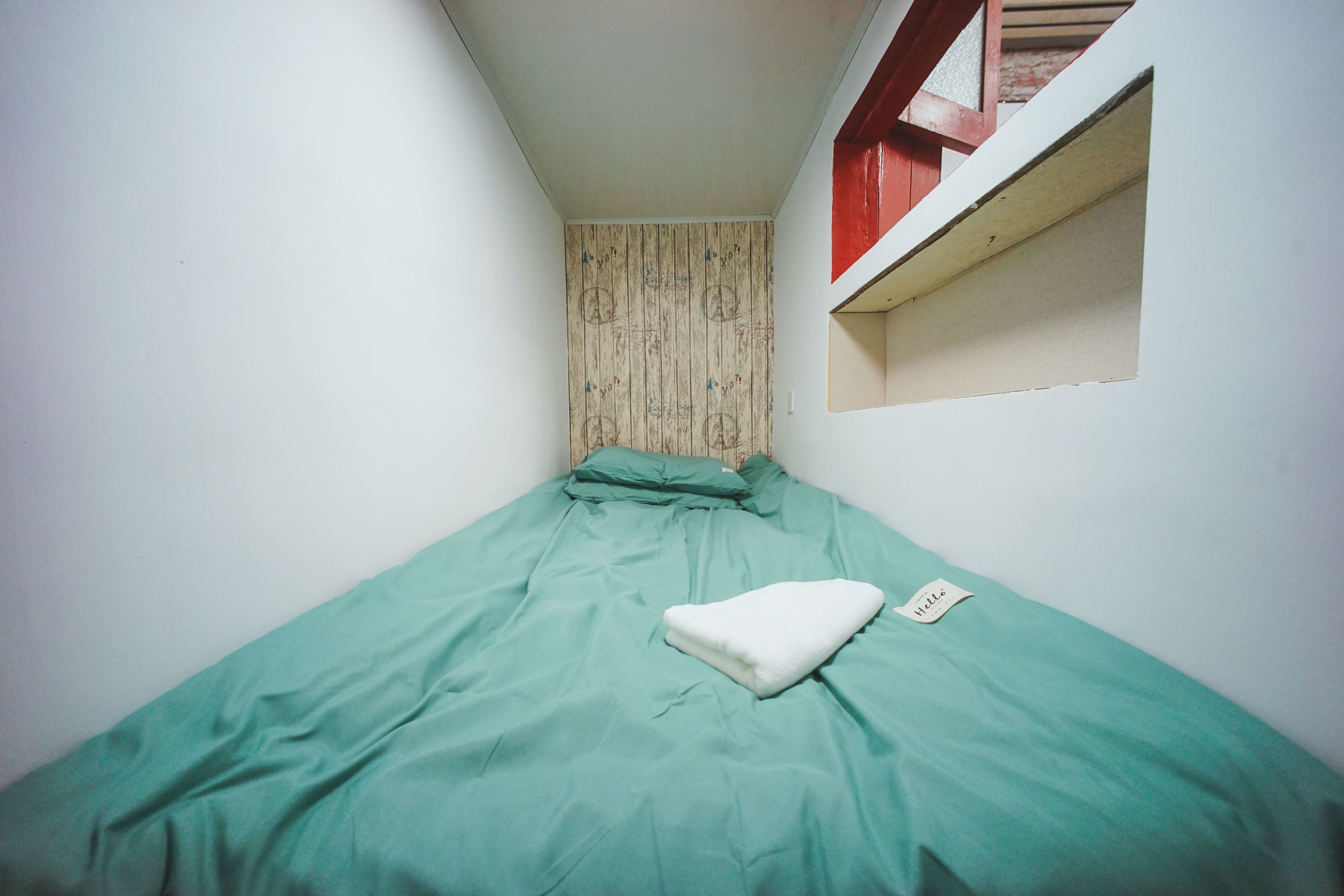A small, single-bed room with a green bedspread, a white pillow, and a small window with a red frame.