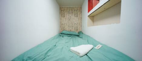 Une petite chambre avec un lit simple, une couvre-lit verte, un oreiller blanc et une petite fenêtre à cadre rouge.