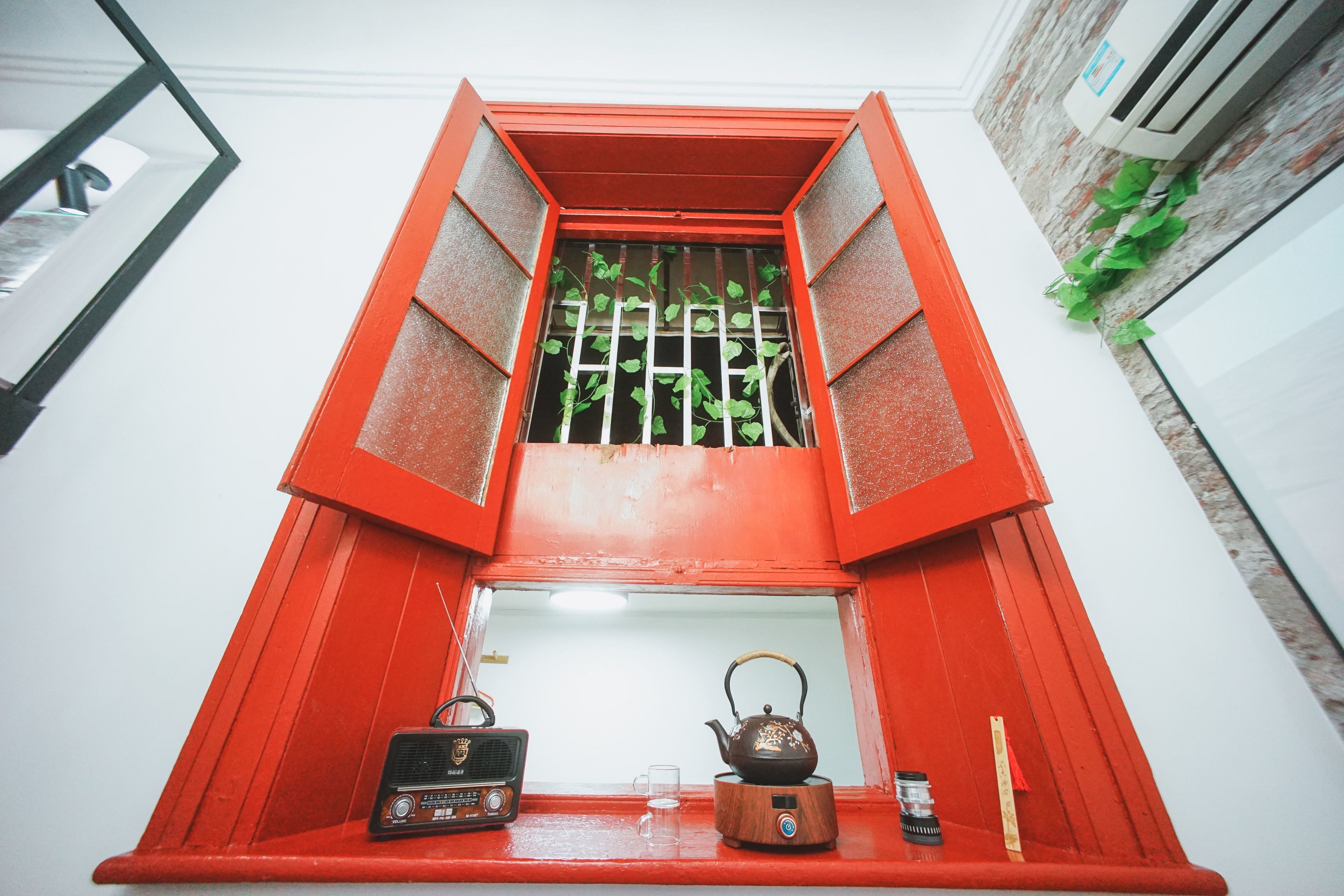A red-framed window with a wooden shelf below, containing a radio, a teapot, and a glass.