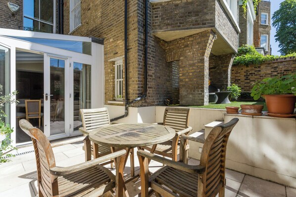An outdoor patio with a round wooden table and six chairs, surrounded by brick walls and potted plants. - Holland Park Avenue VII by Onefinestay (London)