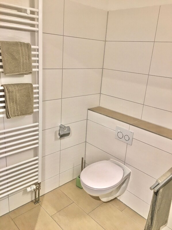 A small, modern bathroom with a toilet, a towel rack with two towels, a radiator, and a green cleaning tool.