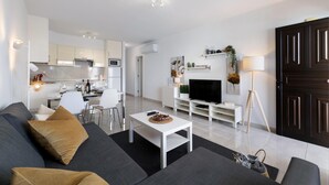 Una sala de estar moderna con un sofá gris, una mesa de centro blanca y un mueble para televisor. Se ve una cocina al fondo con armarios blancos y un microondas.