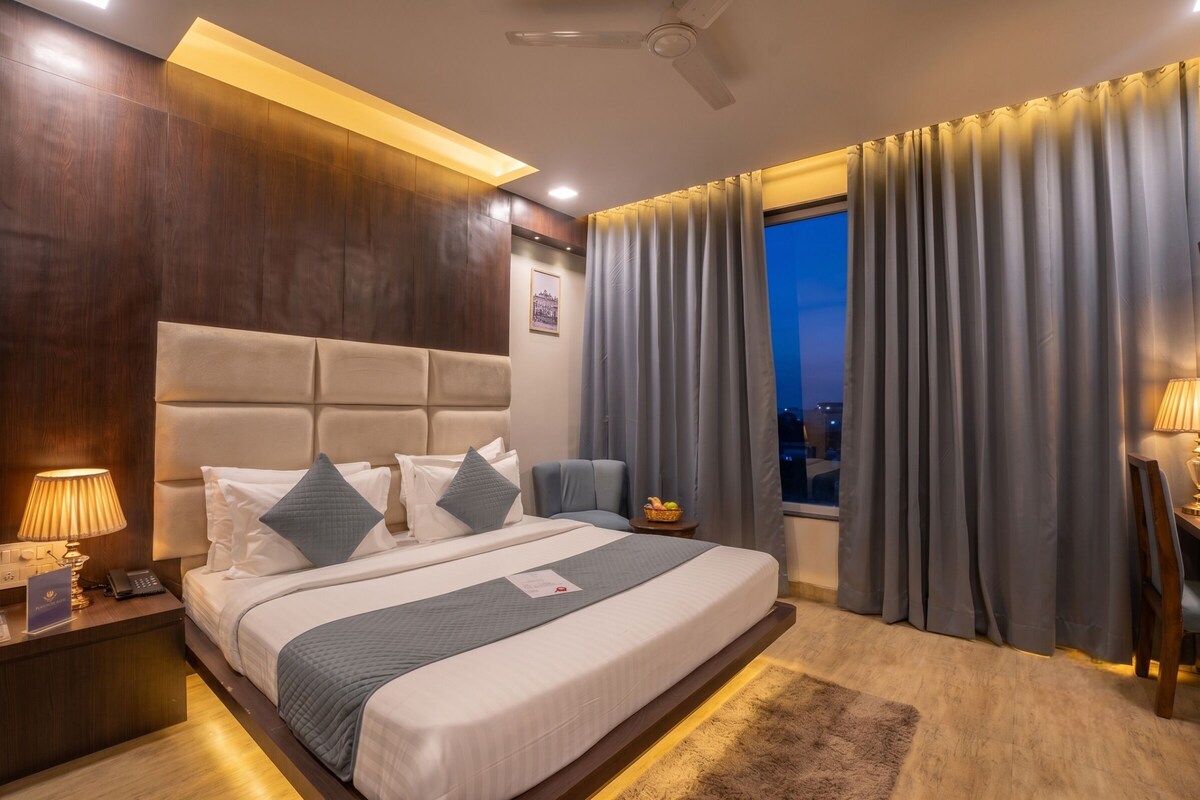 A hotel room with a large bed, a nightstand, a desk, a chair, a window with curtains, and a ceiling fan.