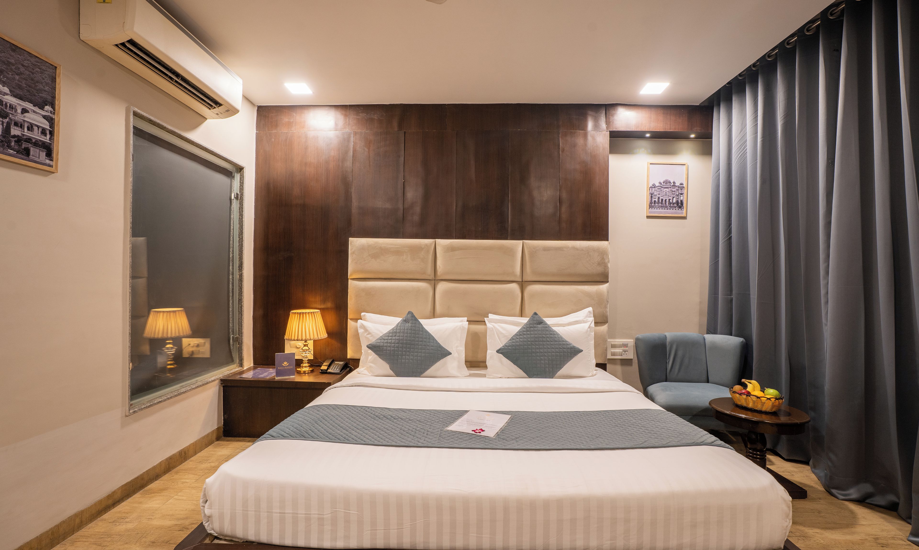 A hotel room with a large bed, a bedside table with a lamp, a chair, and a small table with fruit.