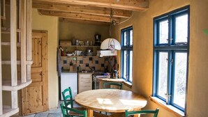 A cozy kitchen with wooden cabinets, a round wooden table, and chairs. There are windows with blue frames, and a hanging light fixture. - Cozy apartment with colored clay plaster, forest-quiet site (Prerow)