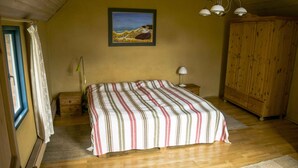 A bedroom with a bed, a wooden wardrobe, a nightstand, a lamp, a painting, and a window with curtains. - Cozy apartment with colored clay plaster, forest-quiet site (Prerow)