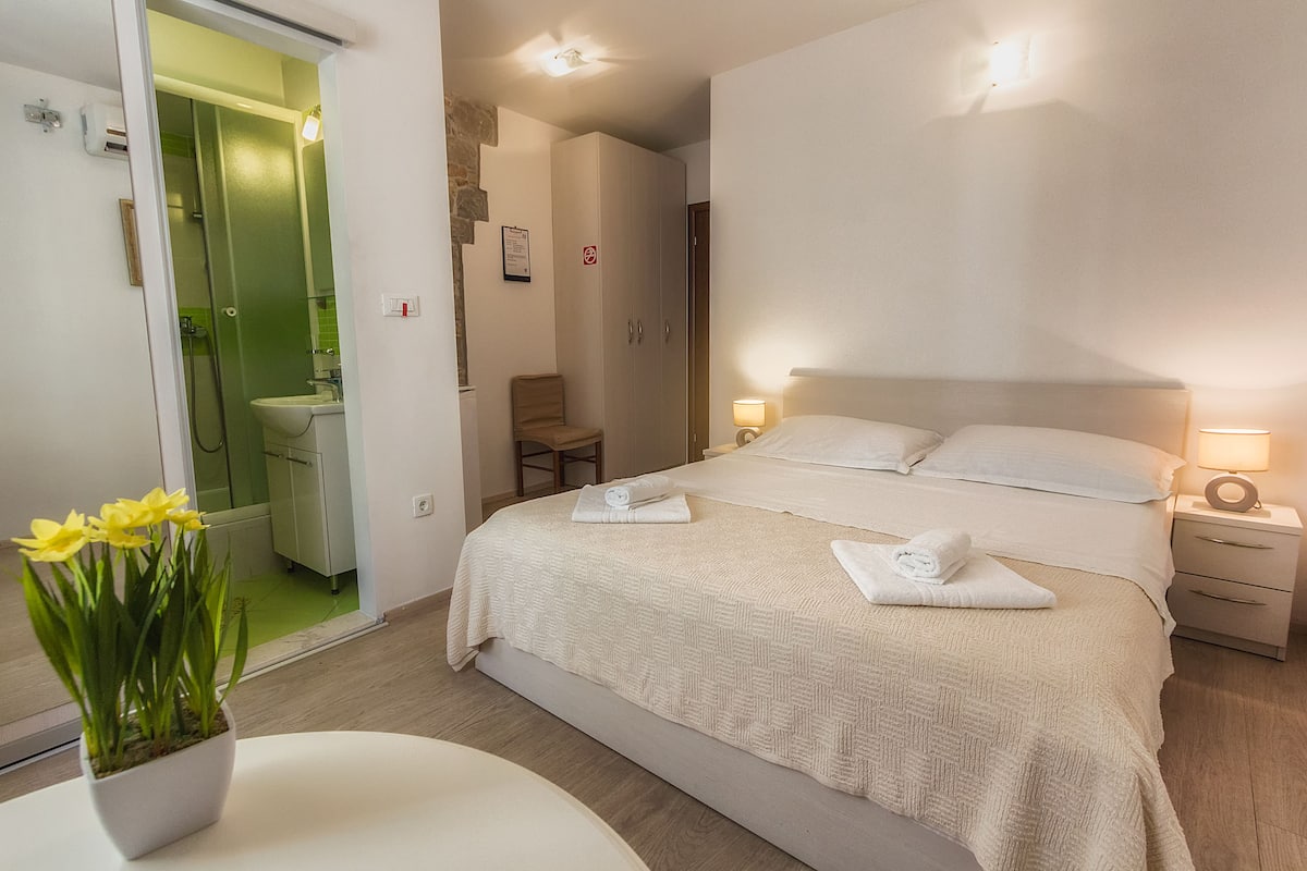 A hotel room with a bed, bedside tables, a chair, a small table with a plant, and a bathroom visible through an open door.