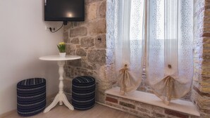 Ett rum med en vägg av sten, en väggmonterad tv, ett litet runt bord med en växt, två randiga sittpuffar och ett fönster med tunna, genomskinliga gardiner.