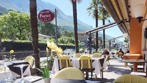 Een terras met eettafels voor twee personen, met helder glaswerk en gele stoelen. Een bord geeft aan dat het Hotel Garni Firenze is.