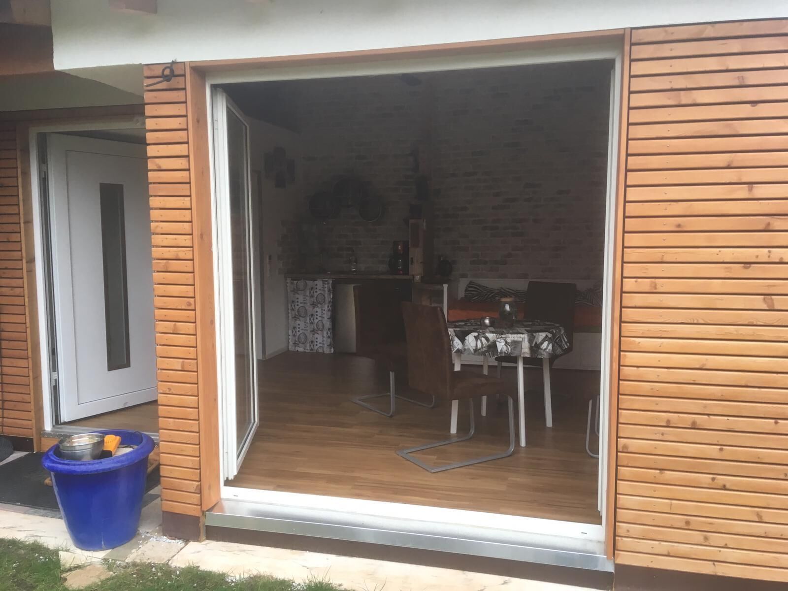 A room with a wooden exterior, a sliding glass door leading to a dining area with a table and chairs, and a blue trash bin outside.
