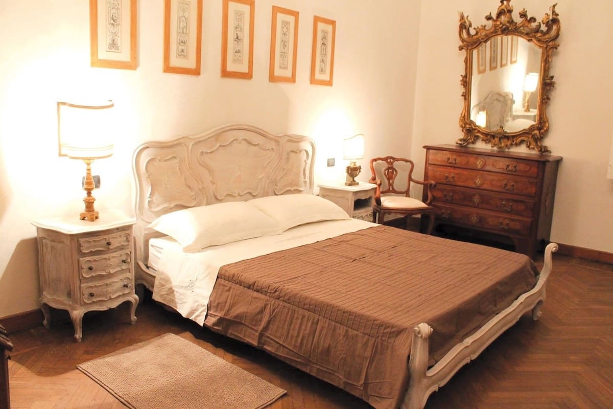A bedroom with a bed, a dresser, a chair, a nightstand with a lamp, and a mirror.