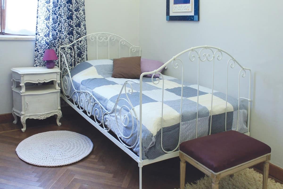 A bedroom with a metal bed frame, a bedside table with a lamp, a round rug, and a framed picture on the wall.