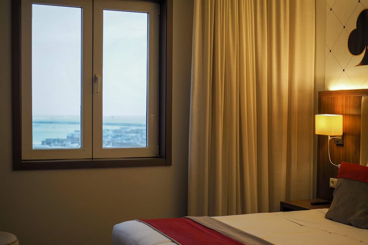 A hotel room with a bed, a nightstand, a lamp, a window with a view, and curtains.