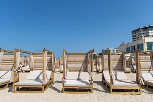 Fila de sillas de playa de madera con cojines color beige, dispuestas sobre una playa arenosa bajo un cielo azul despejado.