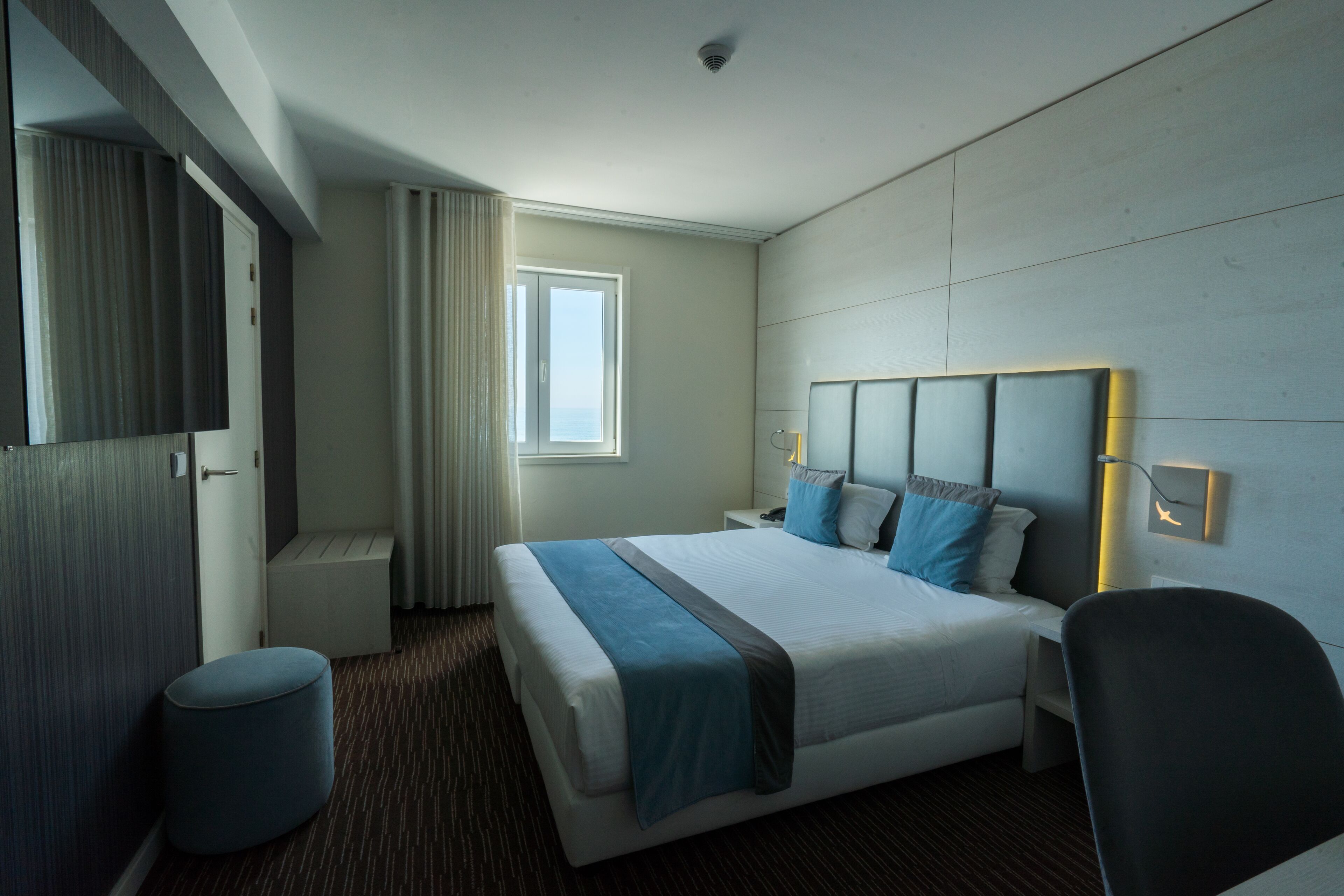 A modern hotel room with a large bed, a bedside table, a chair, a small round ottoman, a window with curtains, and a mirror on the wall.