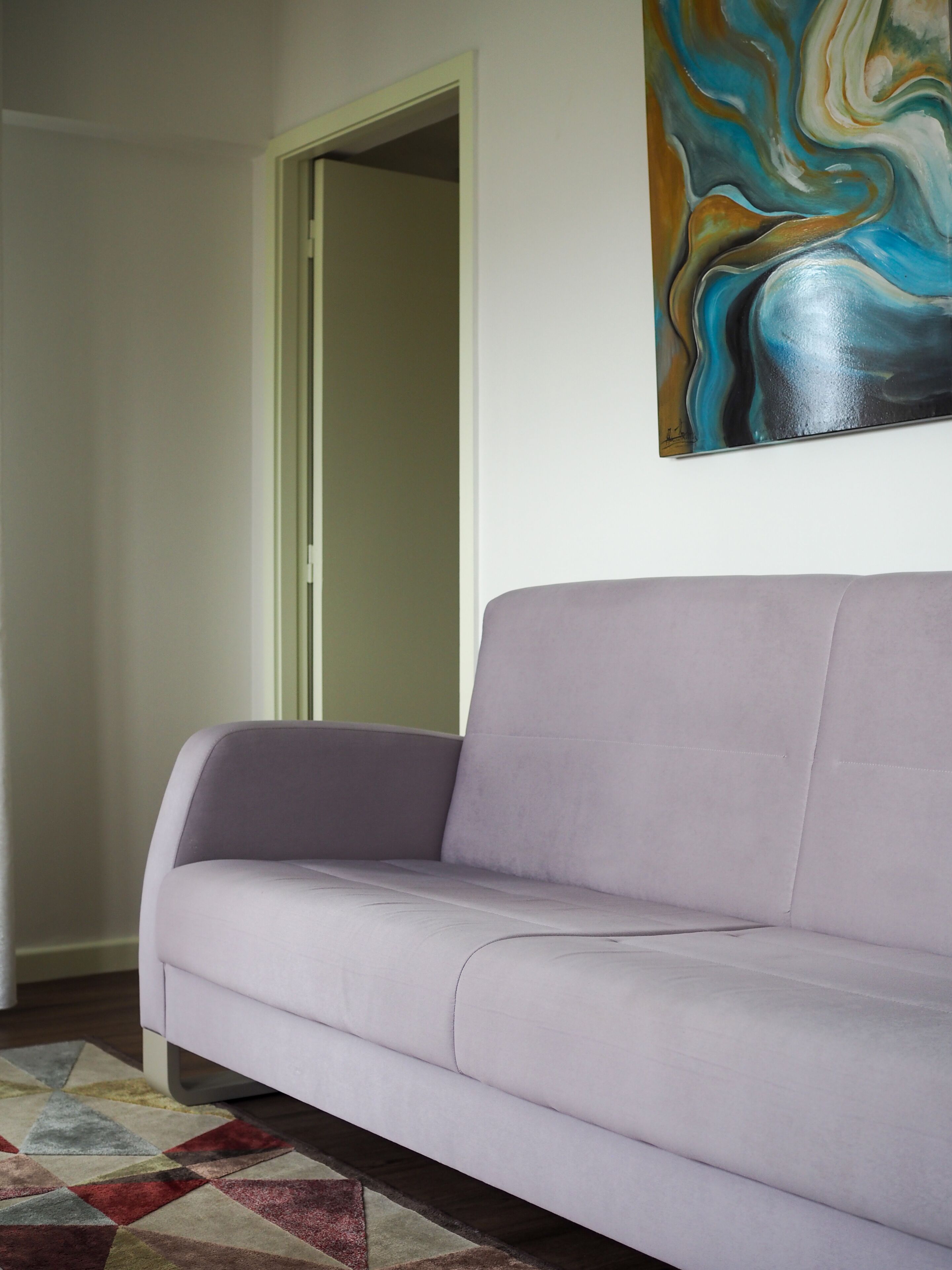 En stue med en lyse lilla sofa, et farverigt abstrakt maleri på væggen og en måtte med geometriske mønstre.