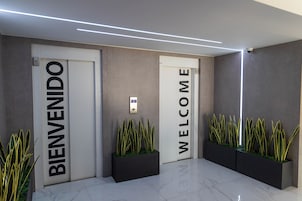 Un vestíbulo moderno con dos puertas, una que dice 'BIENVENIDO' y otra 'WELCOME', flanqueadas por macetas negras con plantas verdes.