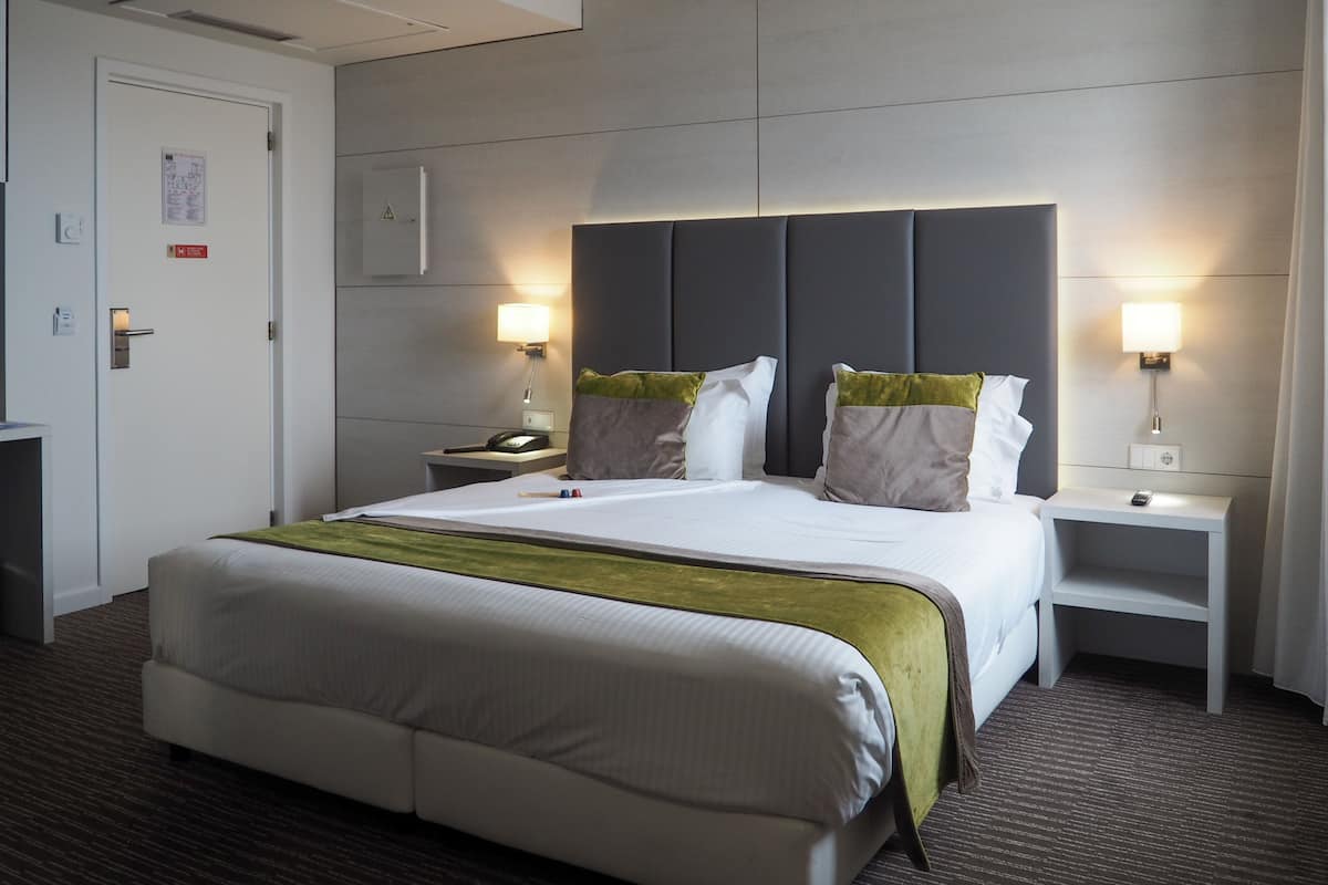 A hotel room with a large bed, two bedside tables, a nightstand, a wall-mounted lamp, and a door with a sign.