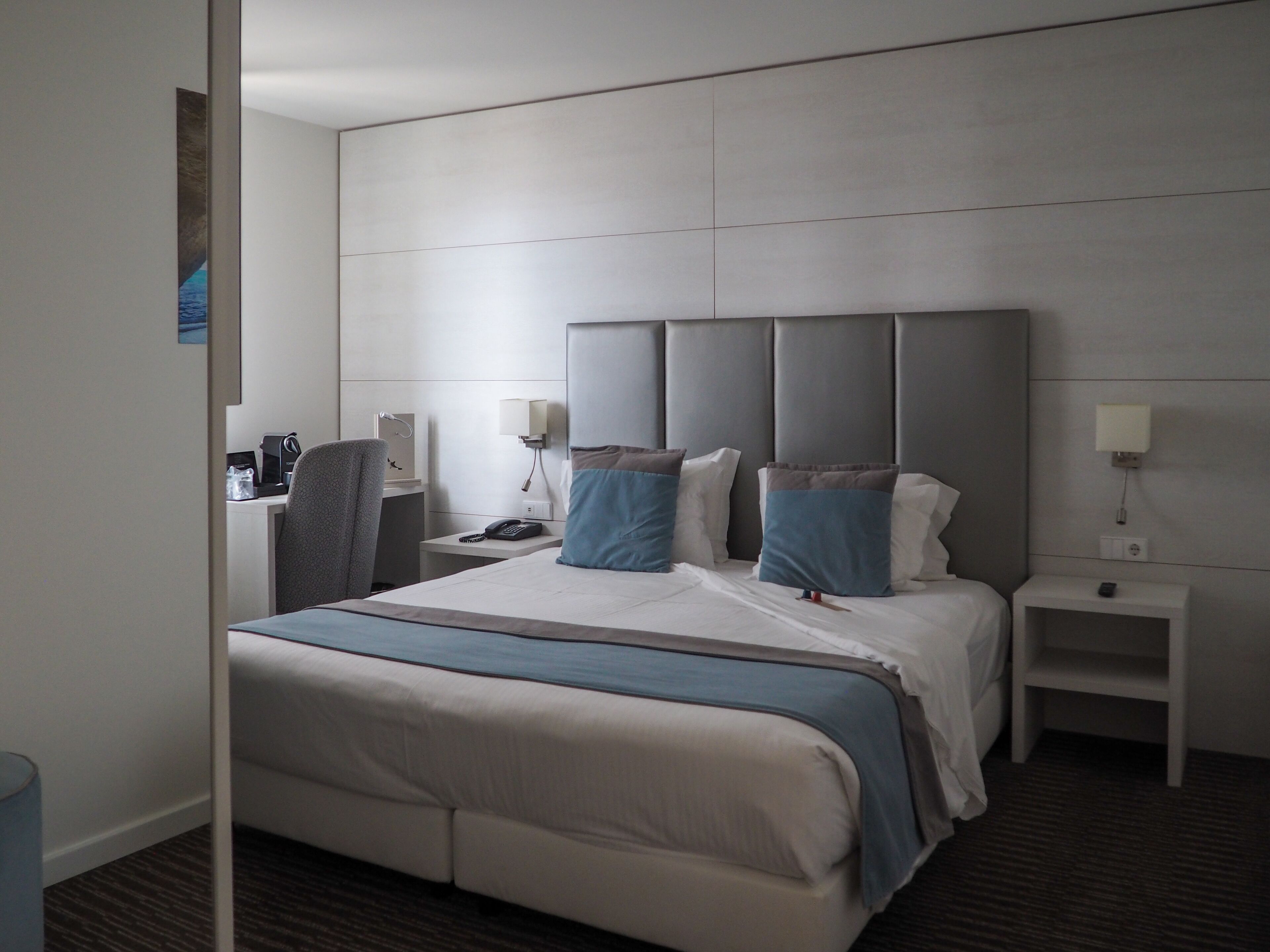 Une chambre d’hôtel moderne, dotée d’un grand lit, d’un bureau et d’une chaise. On y trouve deux tables de chevet avec des lampes et un téléphone.