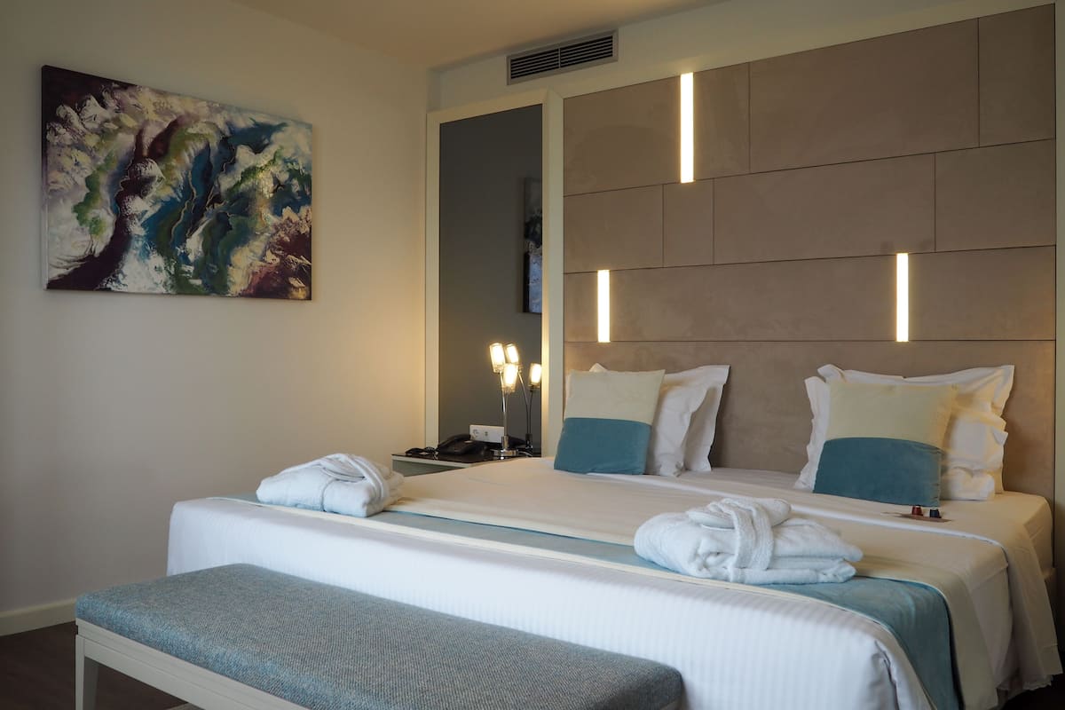 A modern hotel room with a large bed, bedside lamps, a painting on the wall, and a bench.