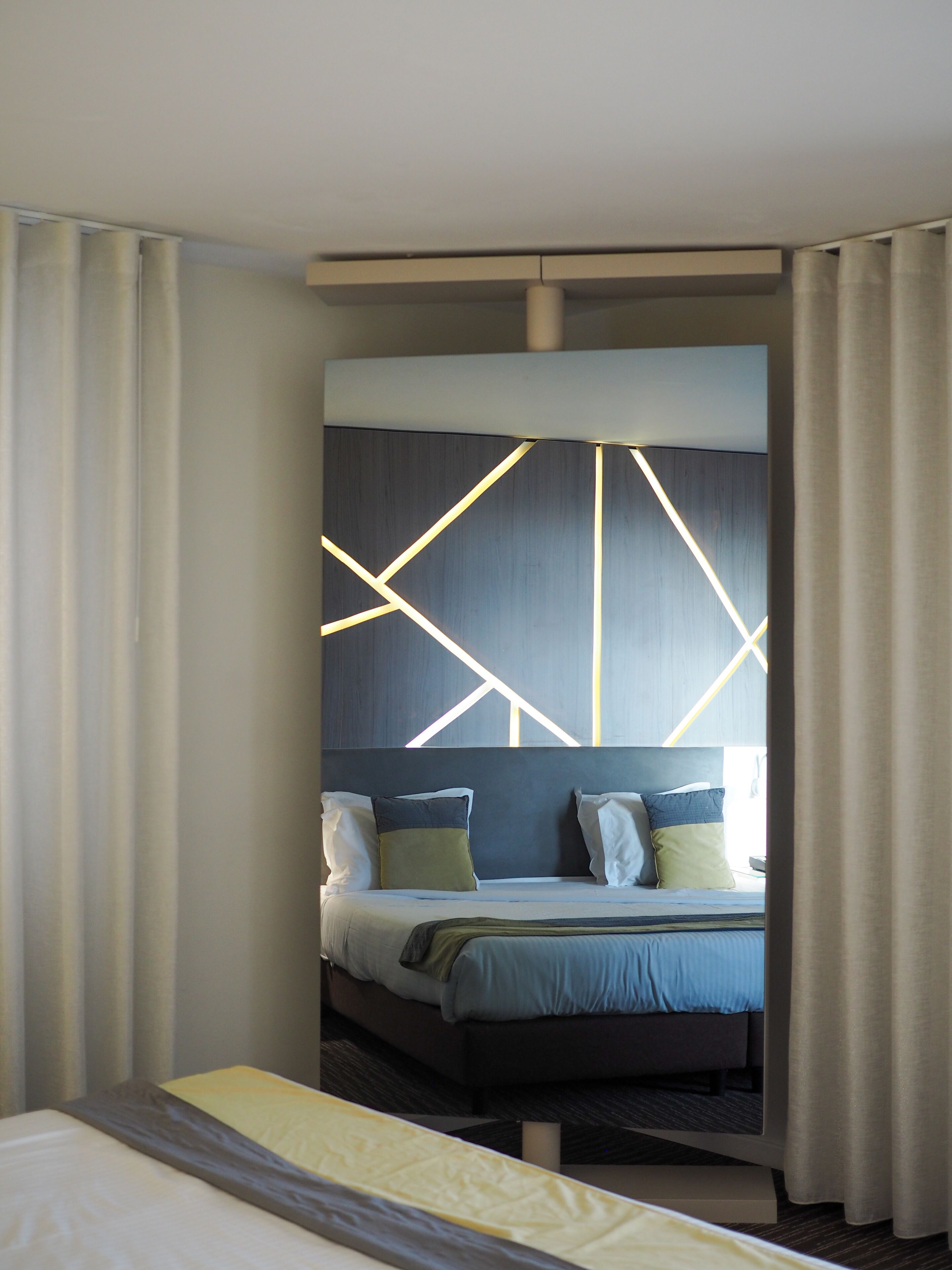 Moderni hotellihuone, jossa on suuri sänky, seinässä geometrisesti muotoiltu valaisin ja näkymä kaupunkiin.