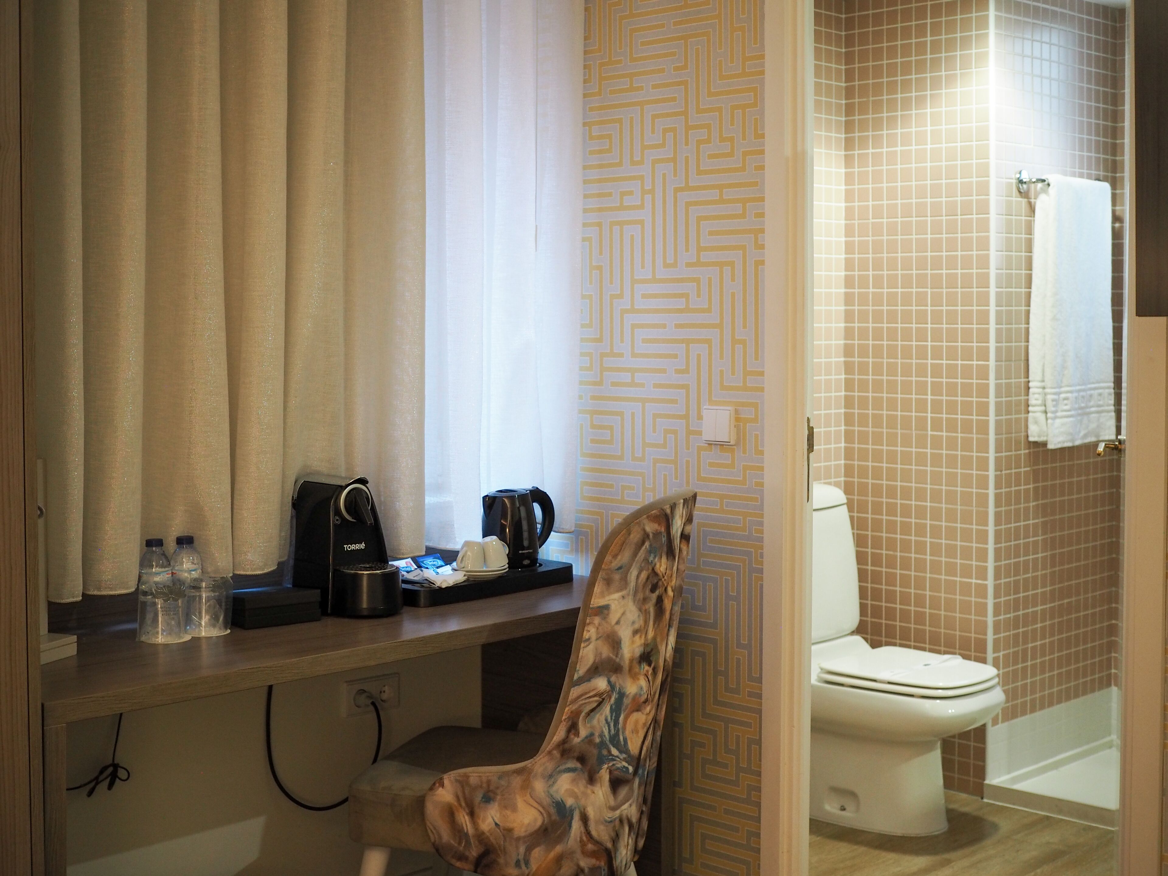 A bathroom with a toilet, a shower, and a sink. There is a chair with a patterned backrest and a towel hanging on the wall.