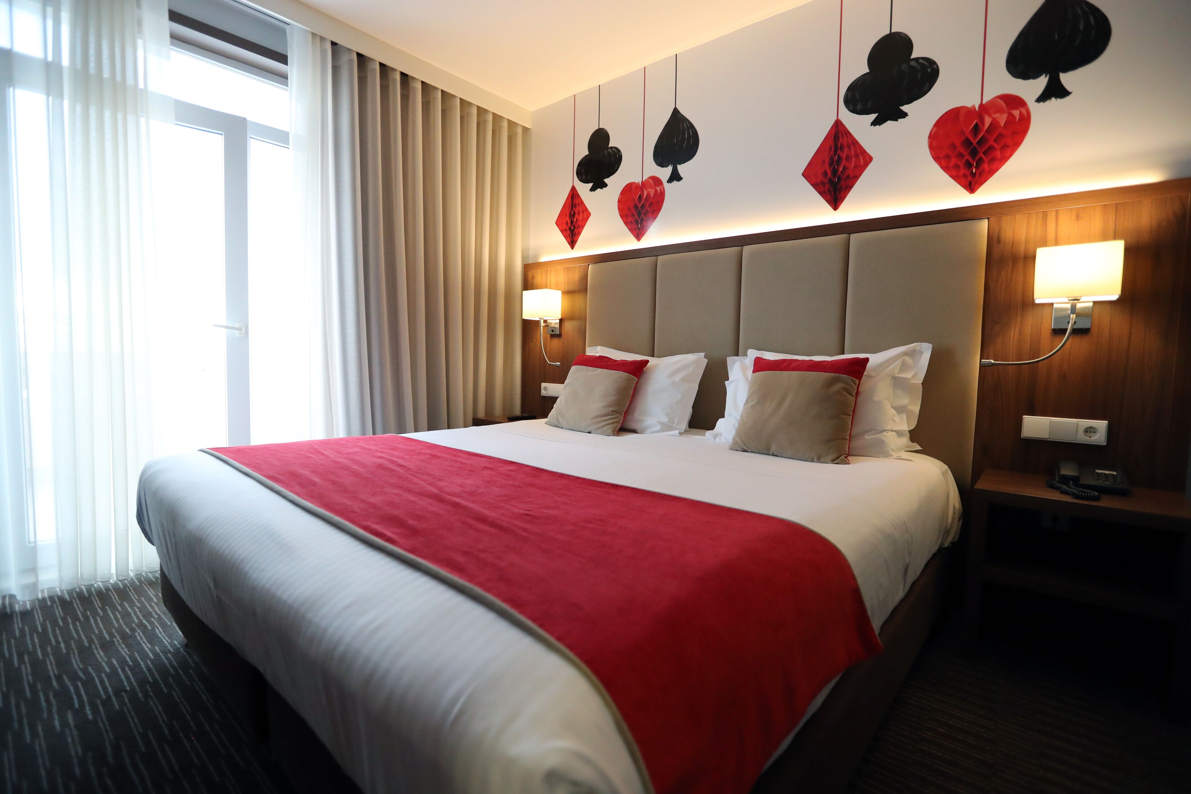 Habitación de hotel con una cama grande, colcha roja y blanca, cabecera decorada con diseños de naipes, dos lámparas de noche y una ventana con cortinas.