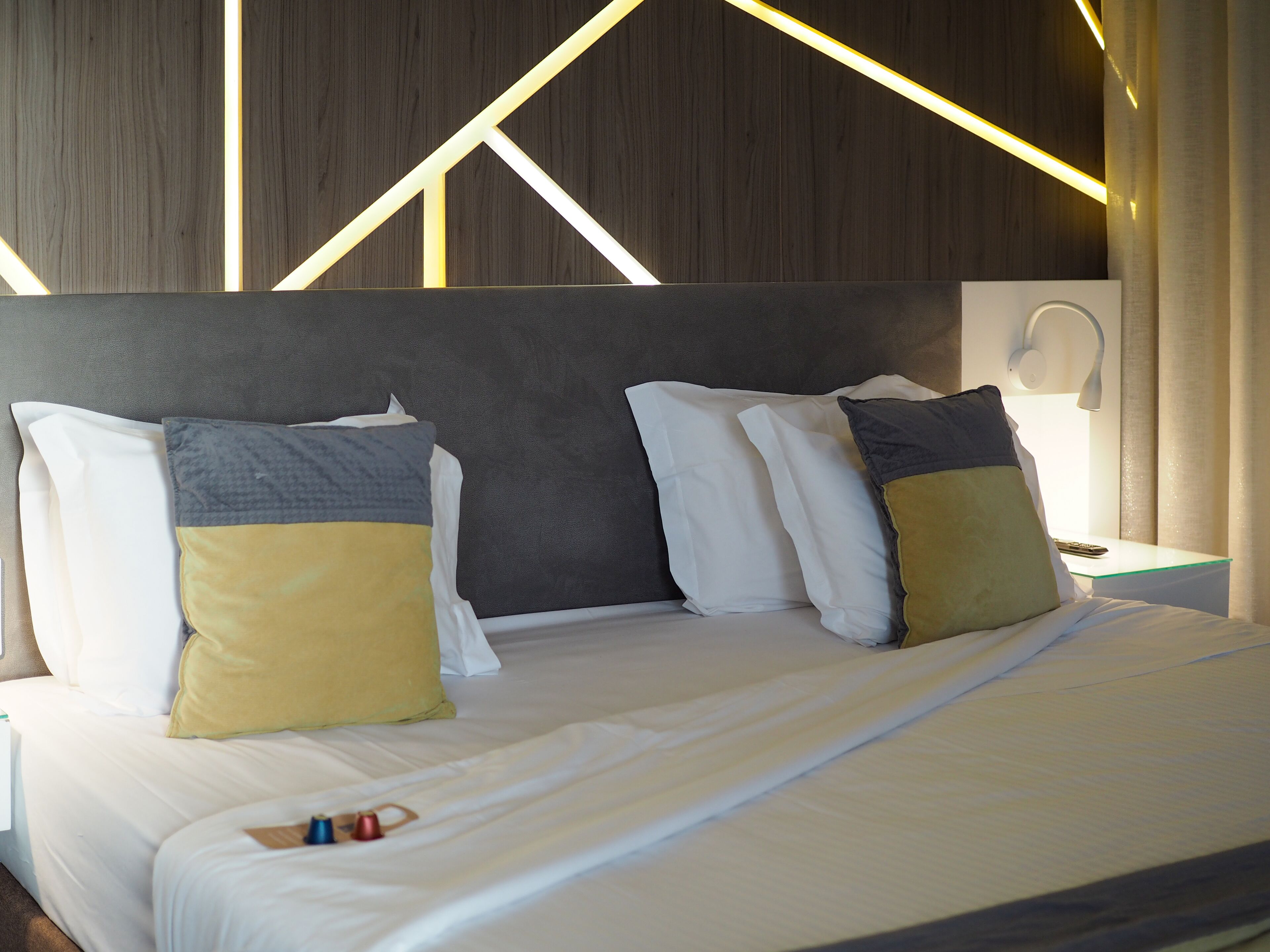 A neatly made bed with white bedding and two pillows, a bedside table with a lamp, and a wall with geometric lighting.