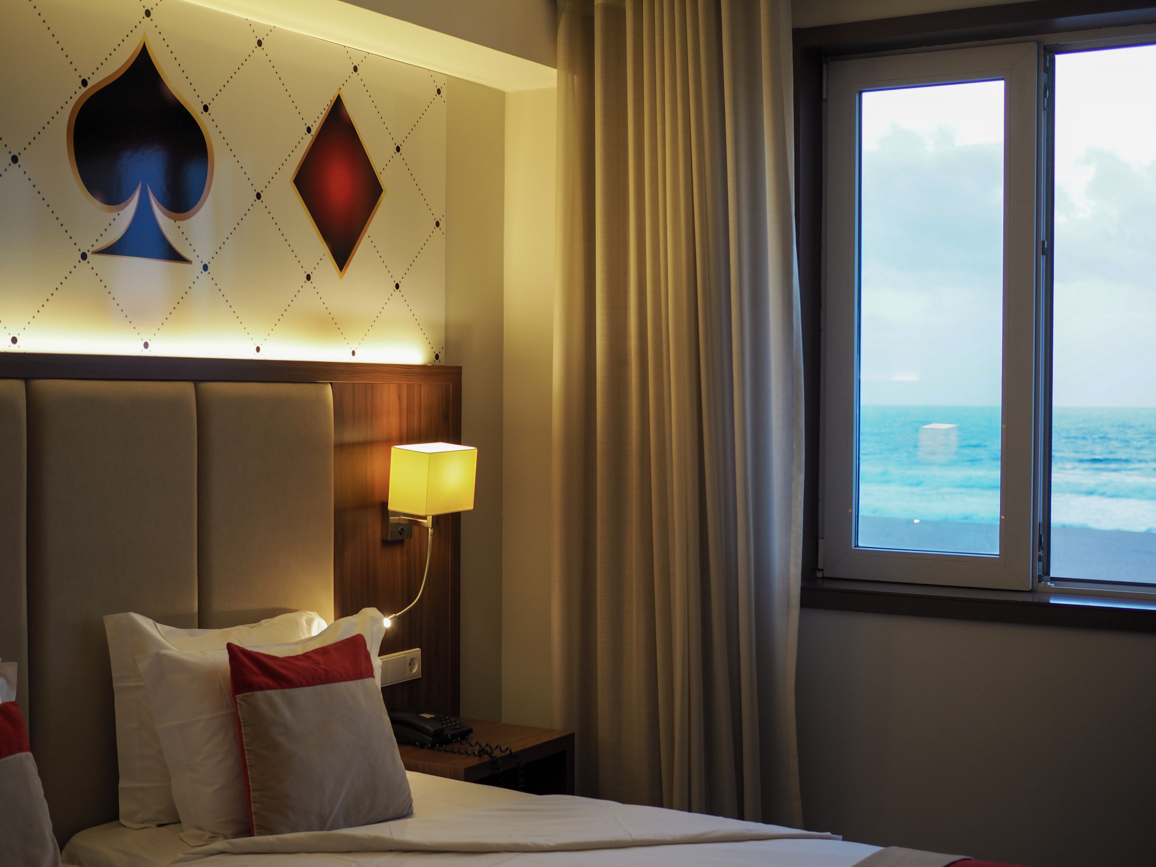 Ein Hotelzimmer mit Bett, Nachttisch mit Lampe, Meerblick durch ein Fenster und eine Wand mit einem Spatel-Symbol.