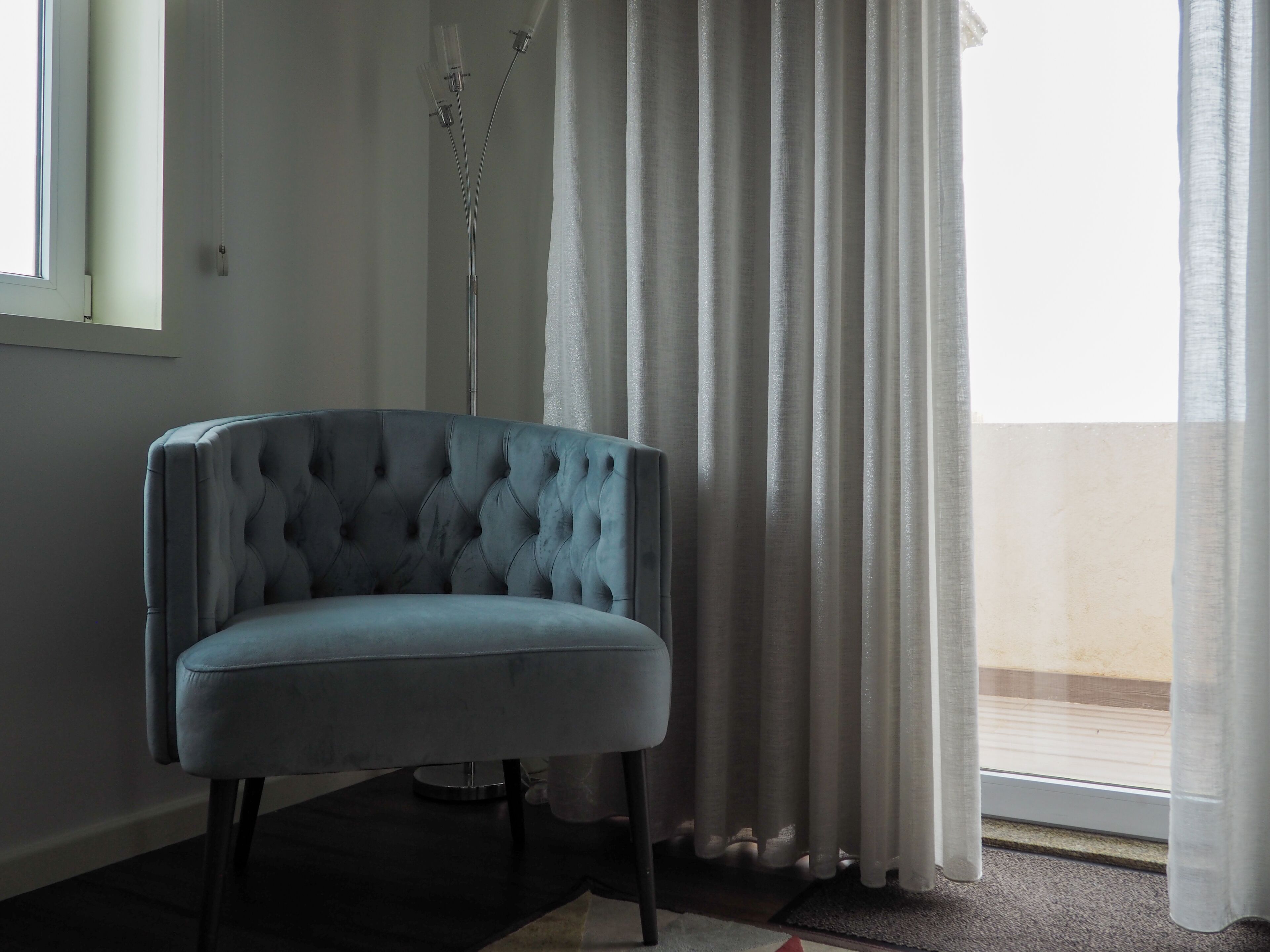 A room with a tufted armchair, a floor lamp, and a view through sheer curtains.