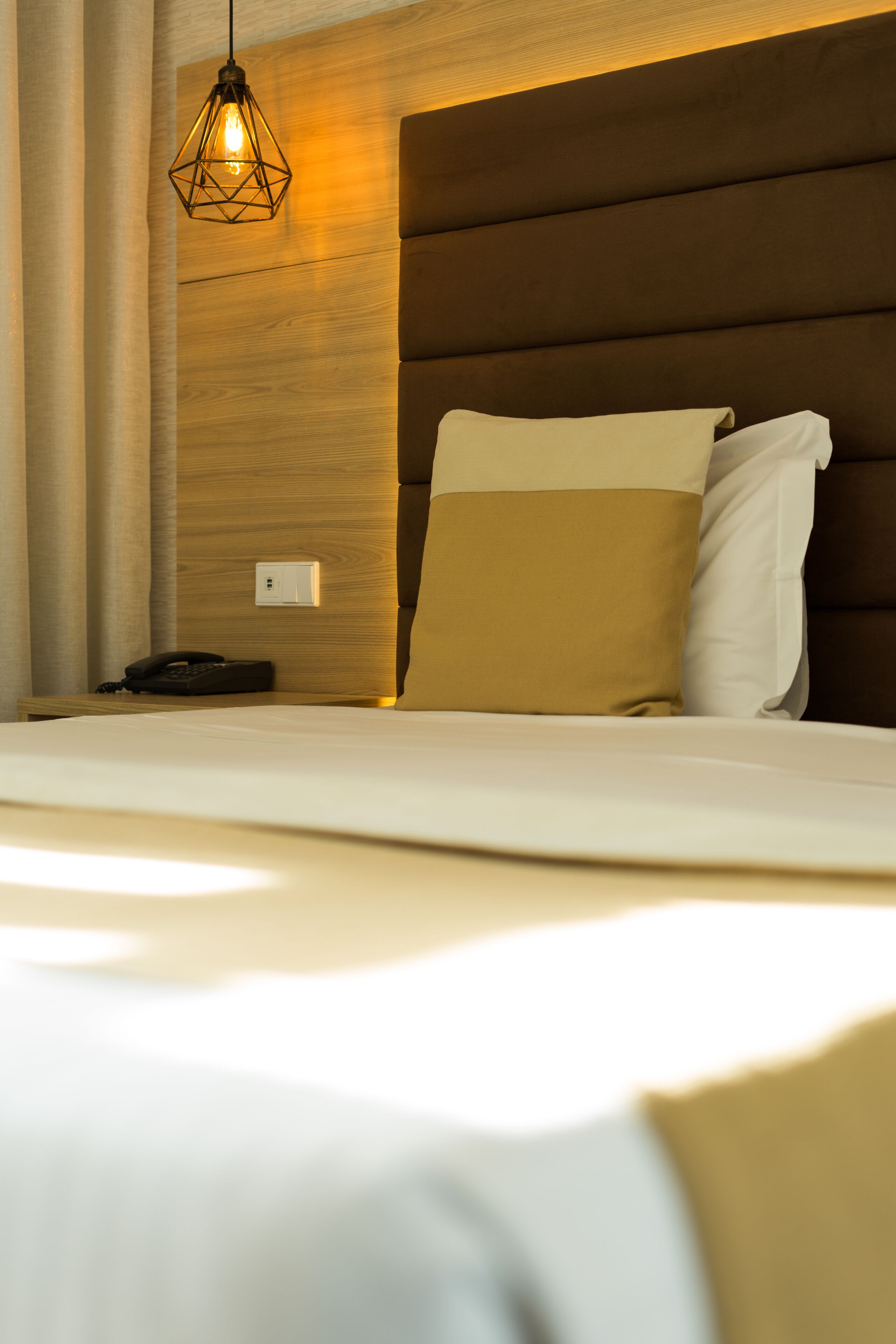 A neatly made bed with a headboard, a bedside telephone, and a hanging light fixture.