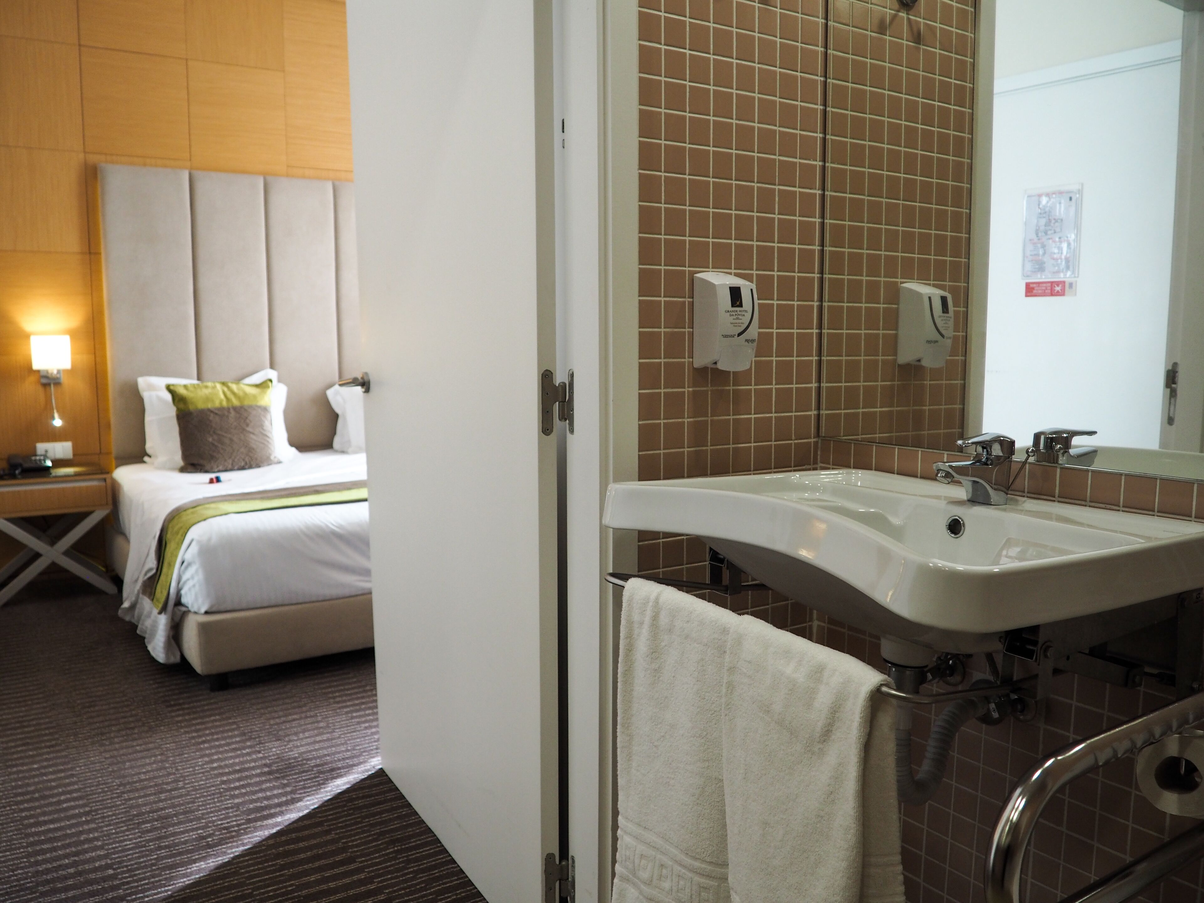 Habitación de hotel con cama, mesita de noche, lámpara y baño con lavabo y espejo.