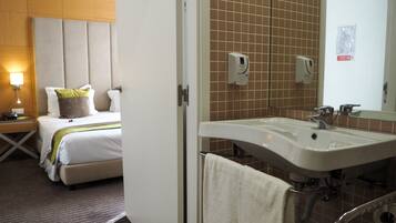 Habitación de hotel con cama, mesita de noche, lámpara y baño con lavabo y espejo.